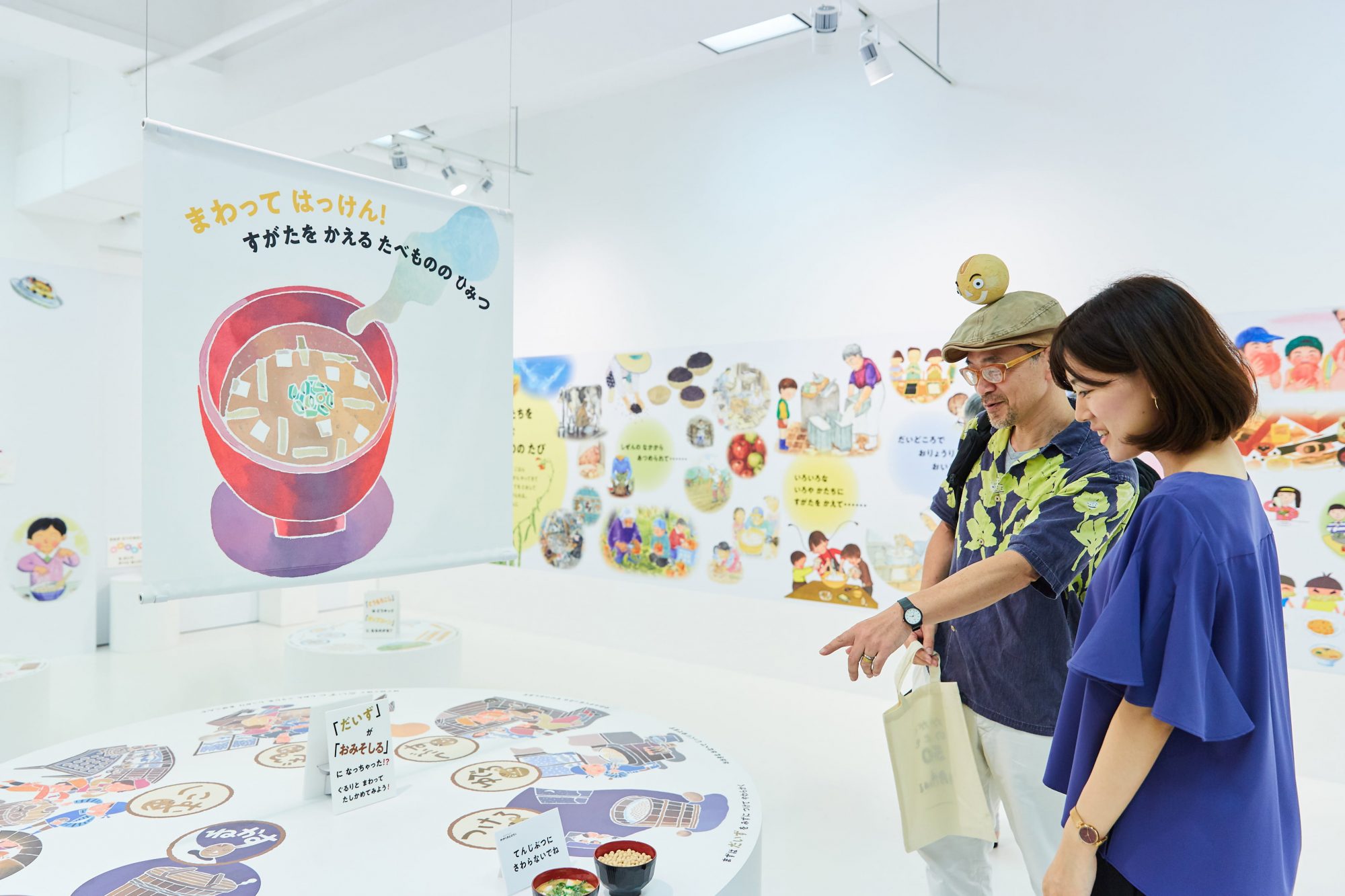 『まんまるダイズみそづくり』の作者であるミノオカ・リョウスケさんと、福音館書店 宣伝課の飛永（とびなが）美紀さんが「たべもの」コーナーで談笑。「601冊の『かがくのとも』には、会場では紹介しきれなかった哲学、工作、仕事といった、たくさんのテーマがあります。今回の展示では『かがくのとも』を知らなかったお子さんや親御さんがいらしたときにも、自分たちの身近にあるものが科学の入口なのだと感じていただけるとうれしいです」（飛永さん）